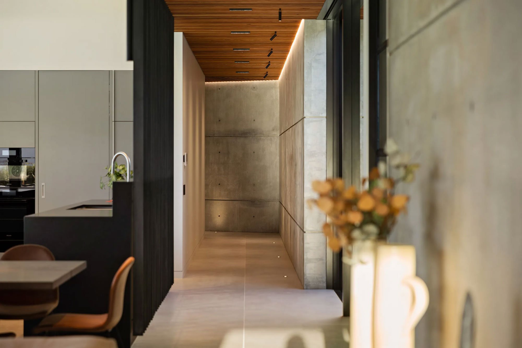 Modern interior hallway with concrete walls, wooden ceiling, contemporary lighting, and a vase of flowers in the foreground.