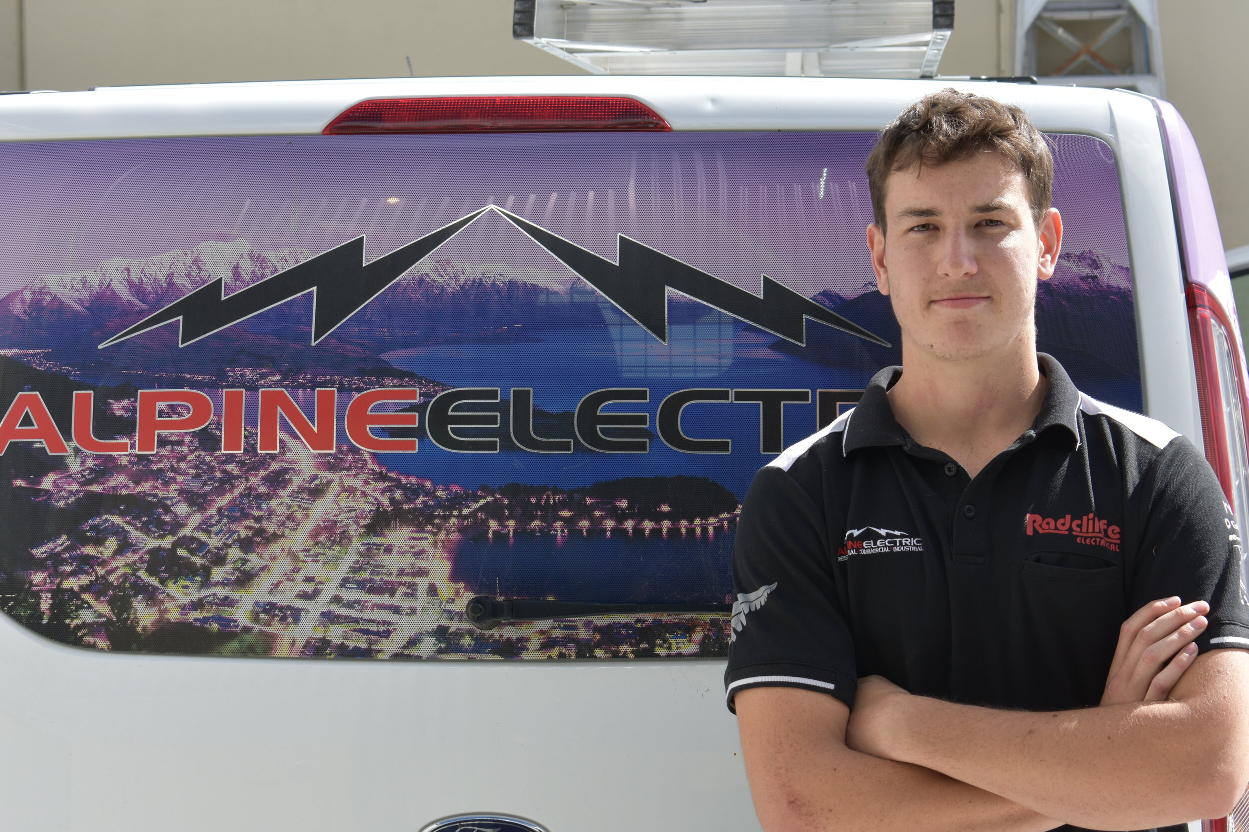 A young man with short brown hair standing with arms crossed in front of a van. The van has a large logo and background image of mountains, a lake, and a cityscape, with the text 'ALPINE ELECTRIC' and lightning bolt graphics.