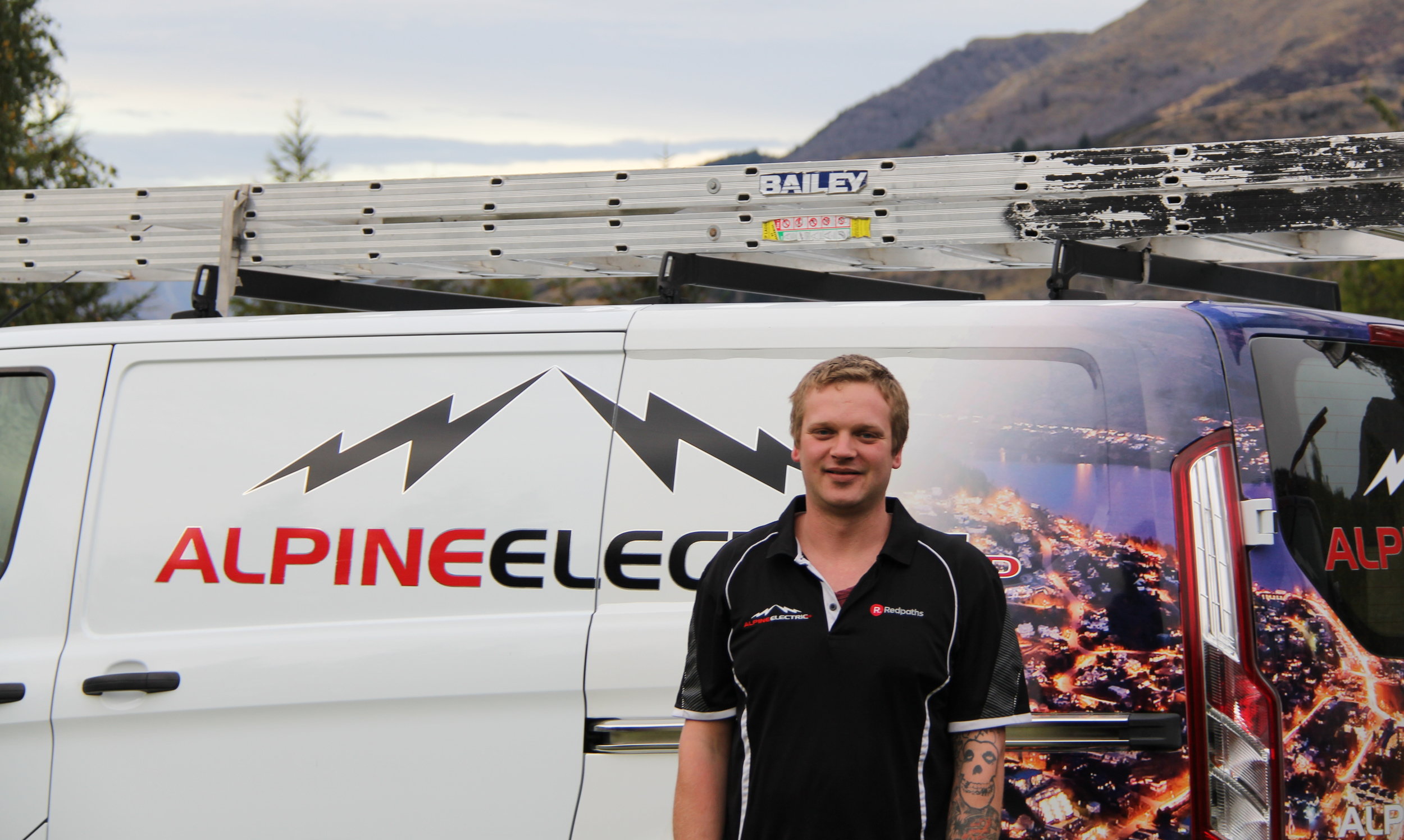 A man standing in front of a white van with the logo 'ALPINE ELECTRIC' and an illustration of mountains and city lights. The man is wearing a black polo shirt with logos and has tattoos on his arm.